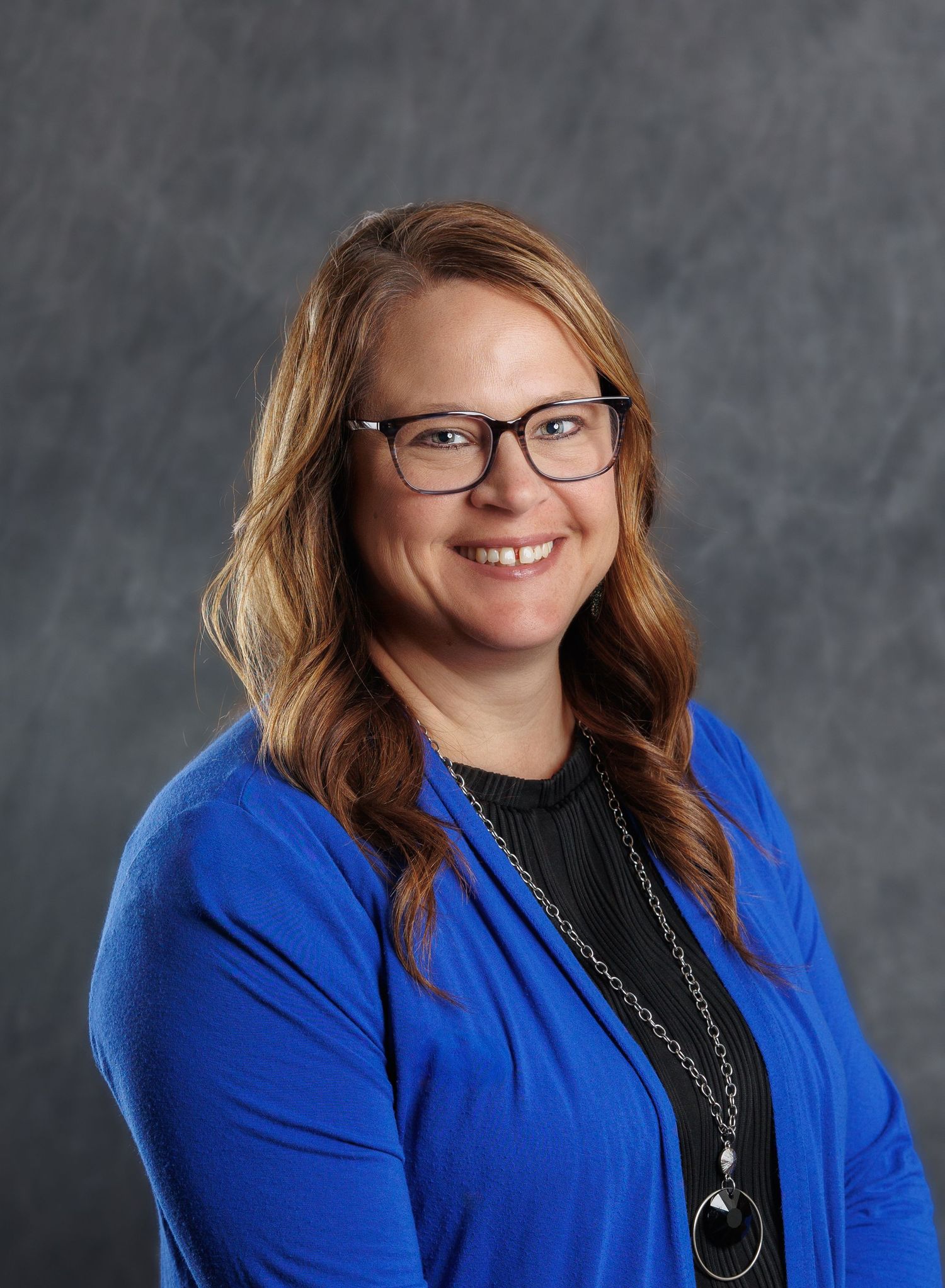 Professional headshot of a person wearing glasses and a royal blue blazer with a black top and necklace against grey backdrop.
