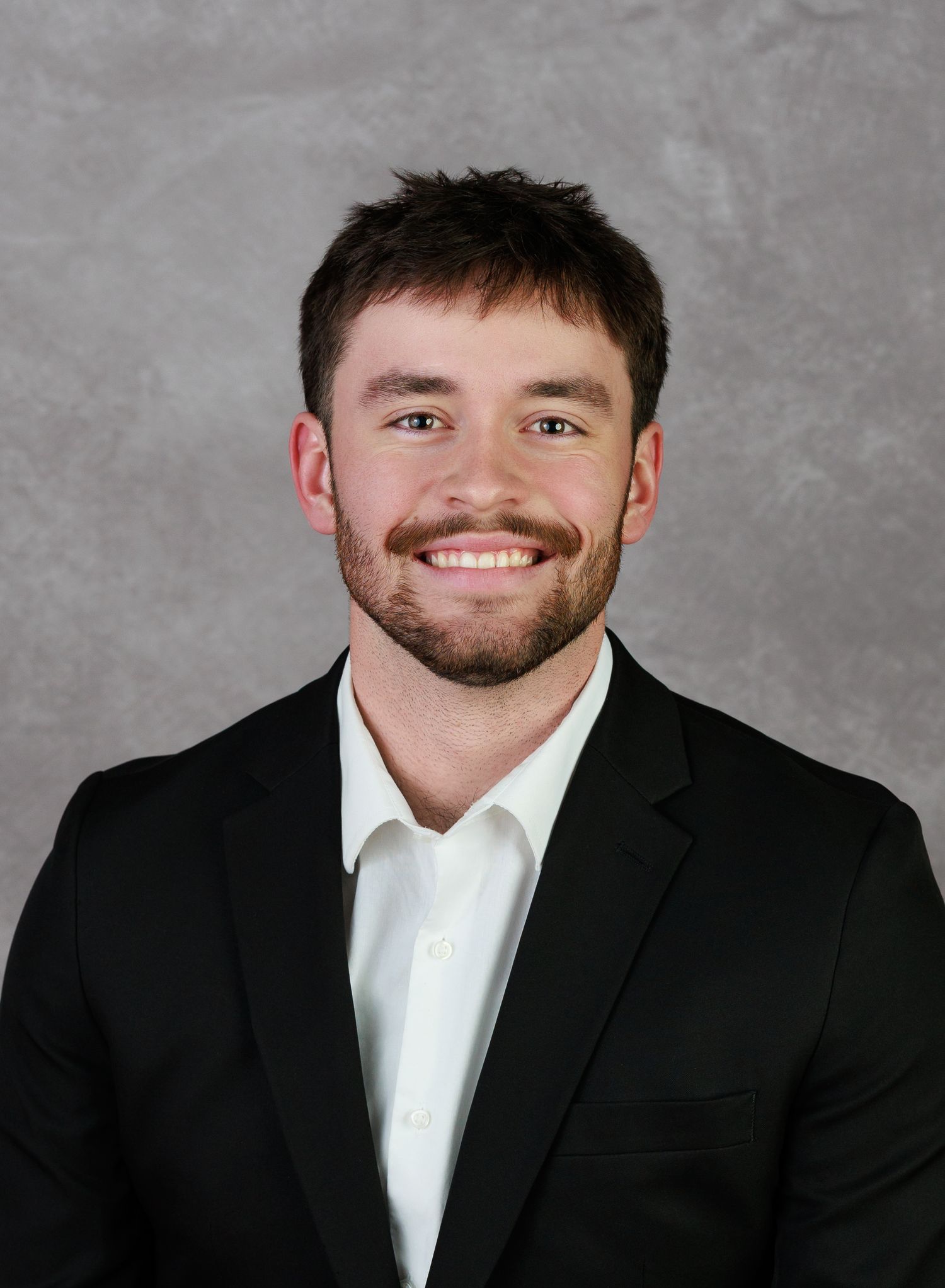 Professional headshot portrait of person in black suit and white shirt against gray background with warm smile.