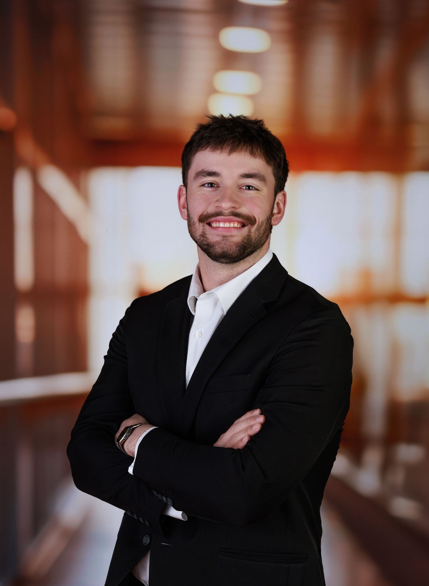 Professional business portrait of someone in a black suit and white shirt smiling warmly against a warm-toned interior background.