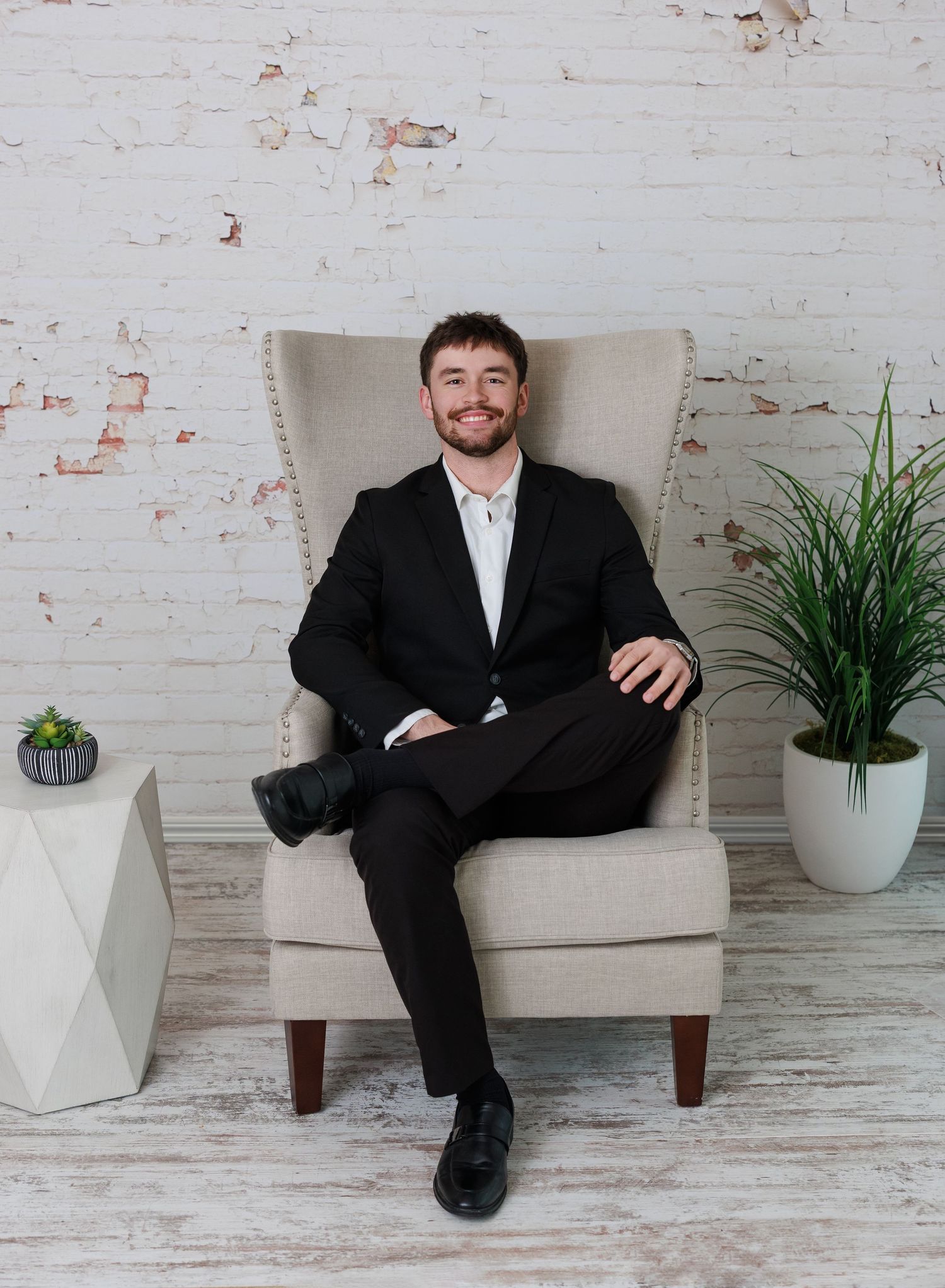 Professional wearing black suit sits casually on modern white chair against rustic brick wall with potted plants.