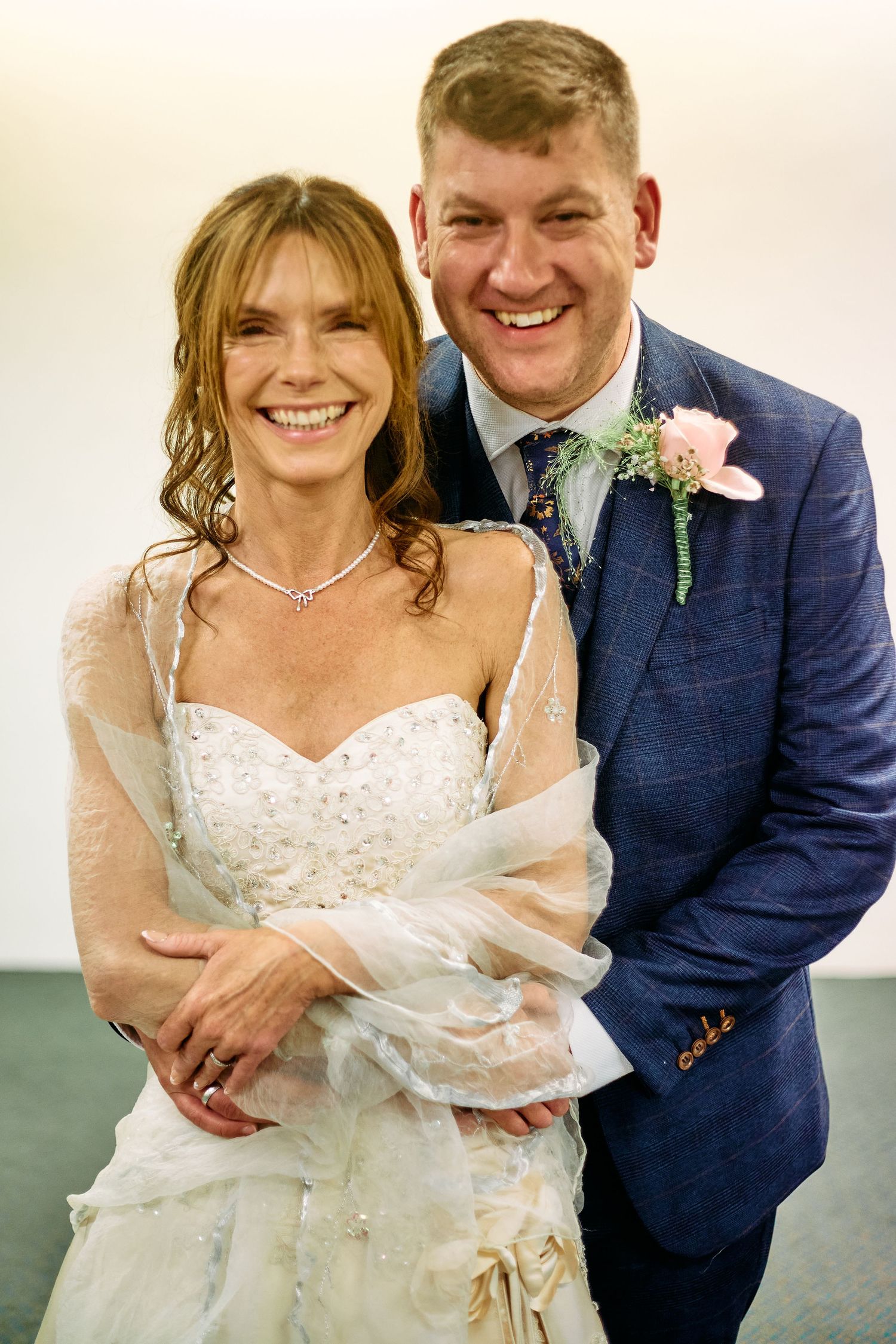 A joyful wedding portrait showing a couple in traditional white dress and navy suit with floral boutonniere.
