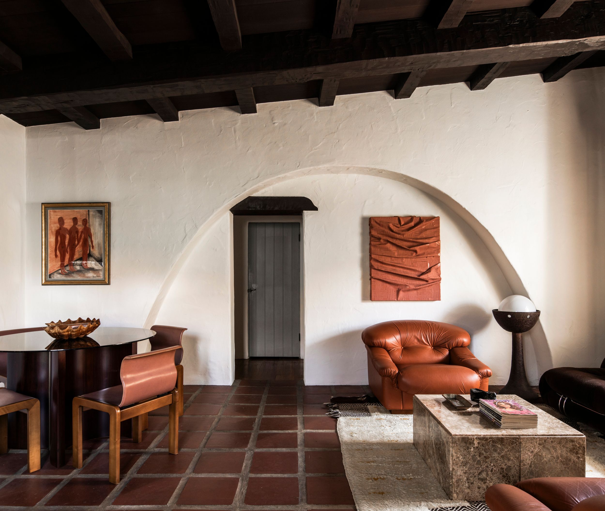 Rustic living room with terracotta tile floors, white arched walls, dark wood beamed ceiling, leather chairs, and abstract artwork.