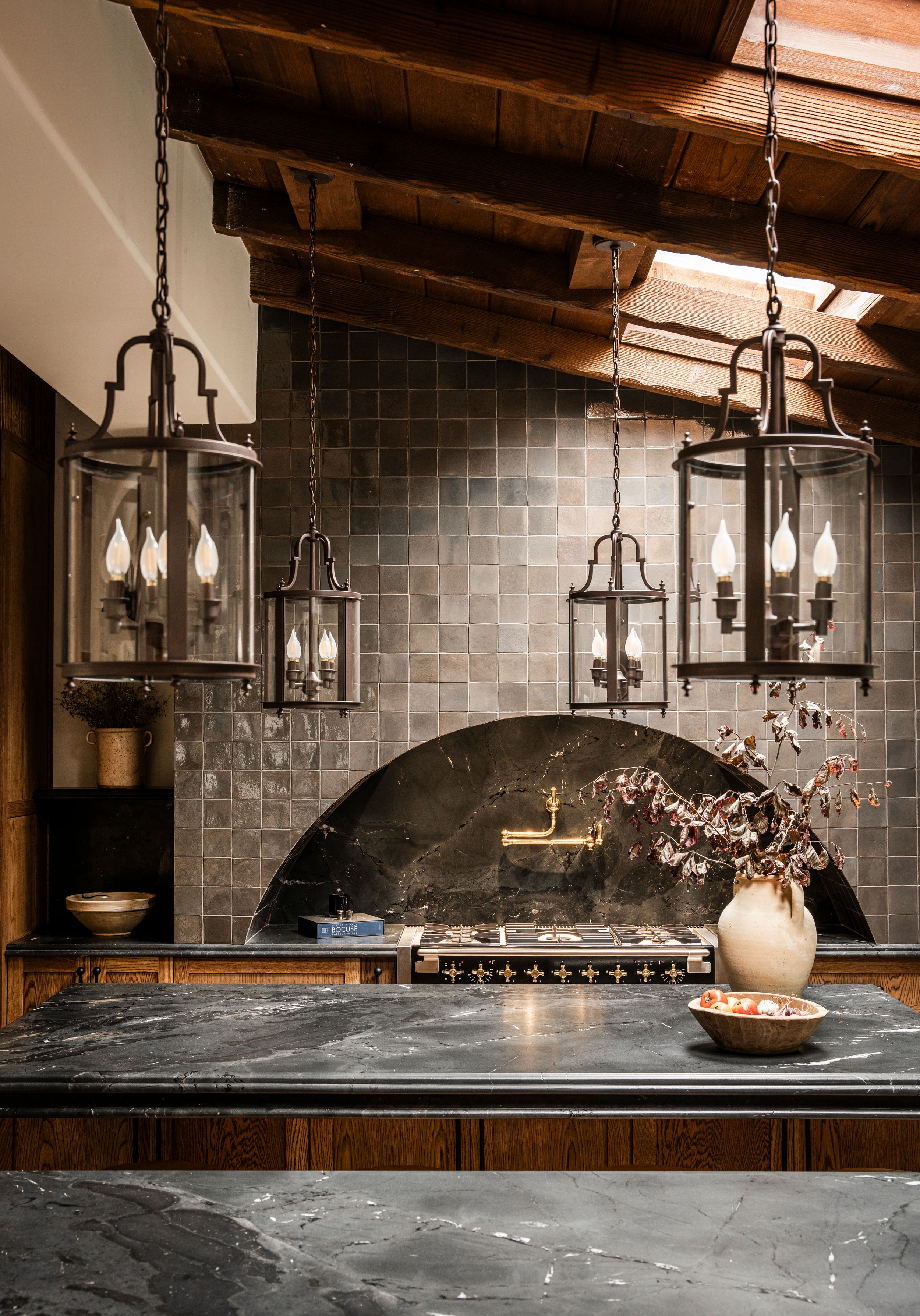 Rustic kitchen interior with hanging lantern pendant lights above dark marble countertop and arched tile backsplash.