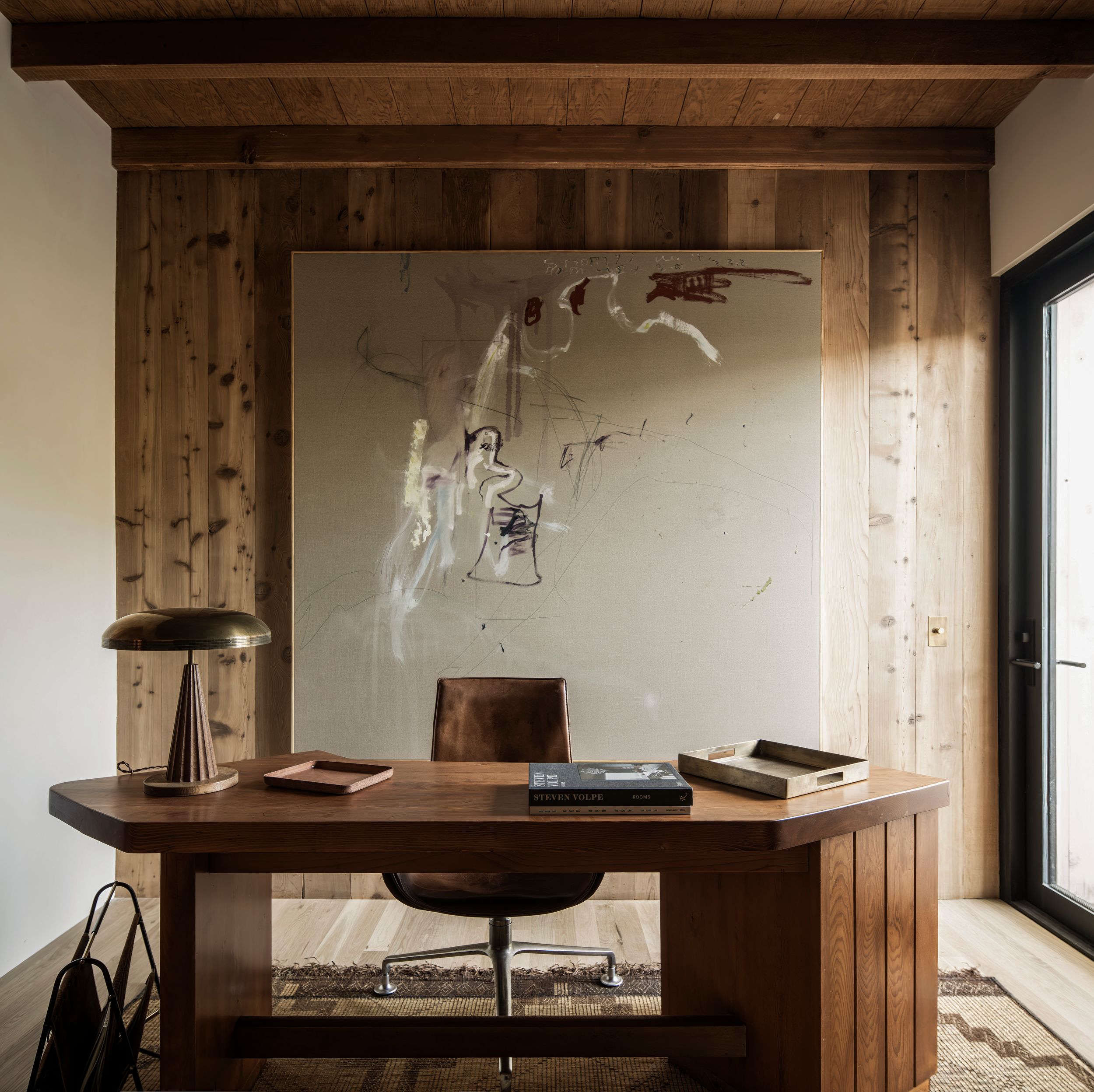 A minimalist wooden desk setup with a table lamp and books against rustic wood paneling and abstract wall art.