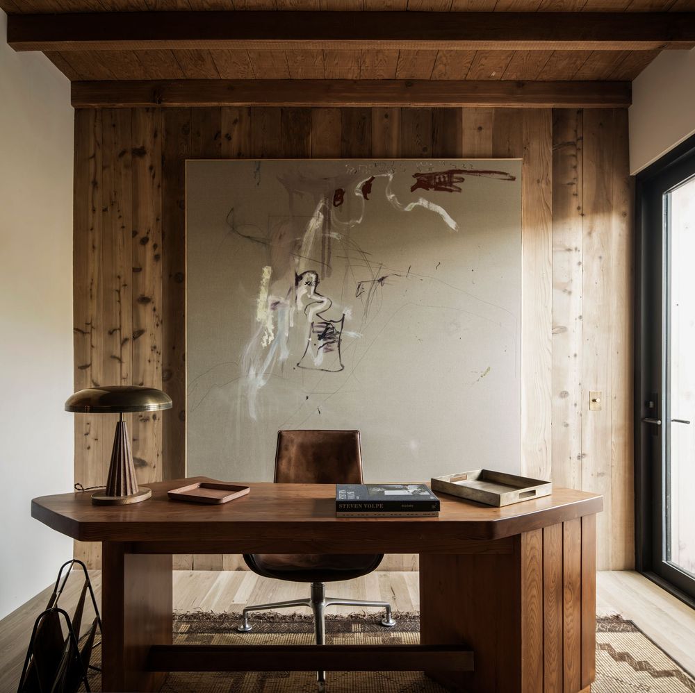 A minimalist wooden desk setup with a table lamp and books against rustic wood paneling and abstract wall art.