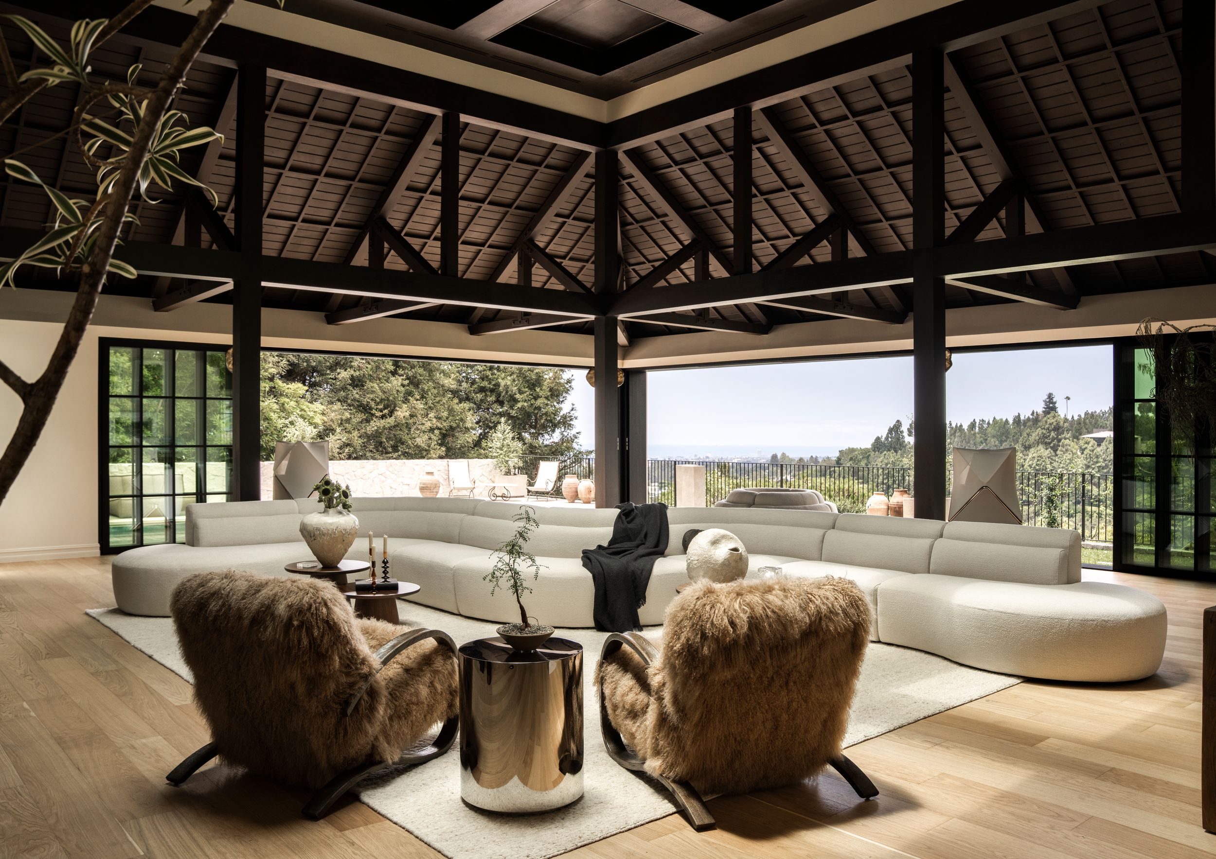 Luxurious open-plan living room with vaulted wooden ceiling, white curved sofa, fur chairs, and panoramic views of lush greenery.