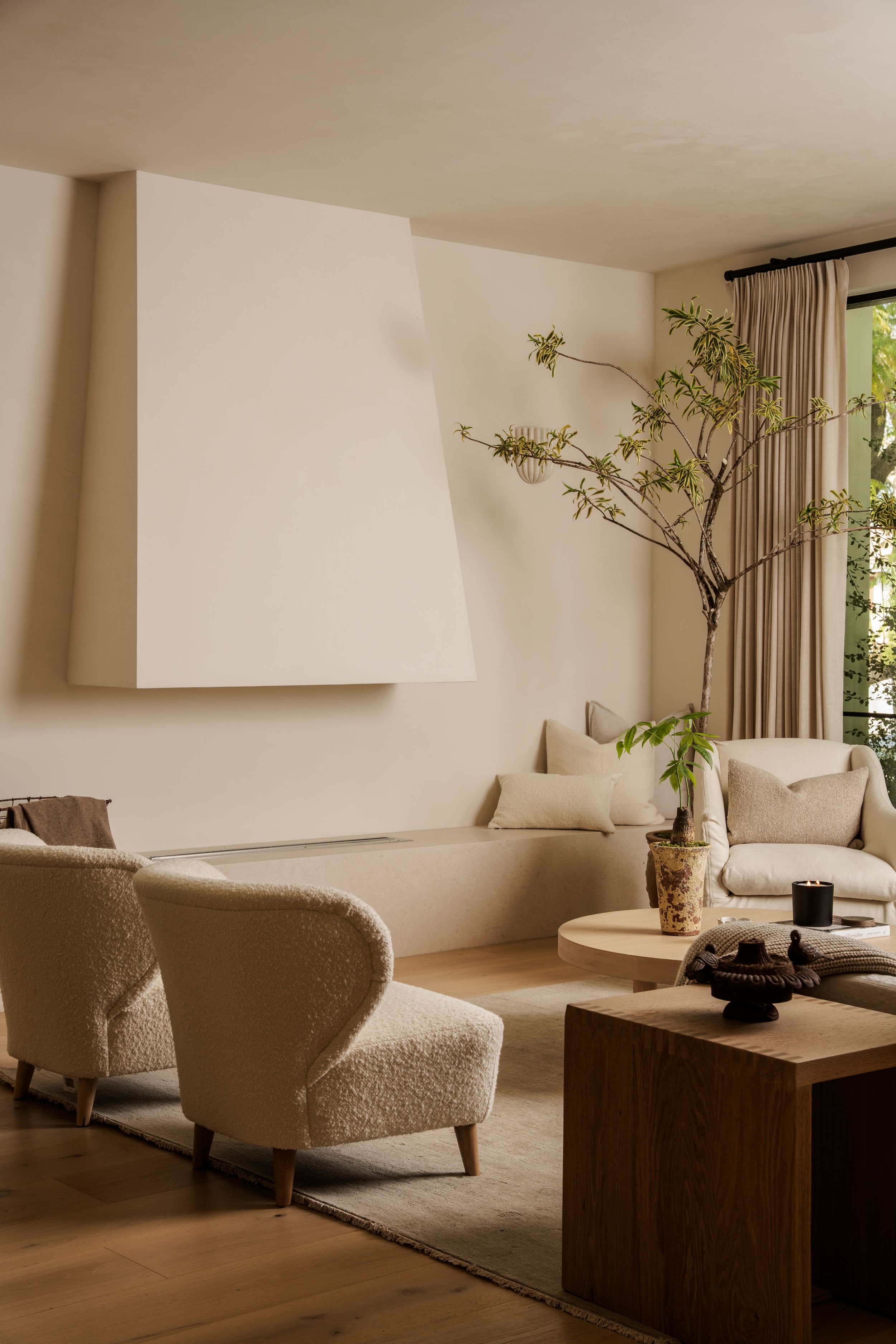 Minimalist living room with beige bouclé armchair, wooden coffee table, neutral sofa, and tall branch in vase by window.