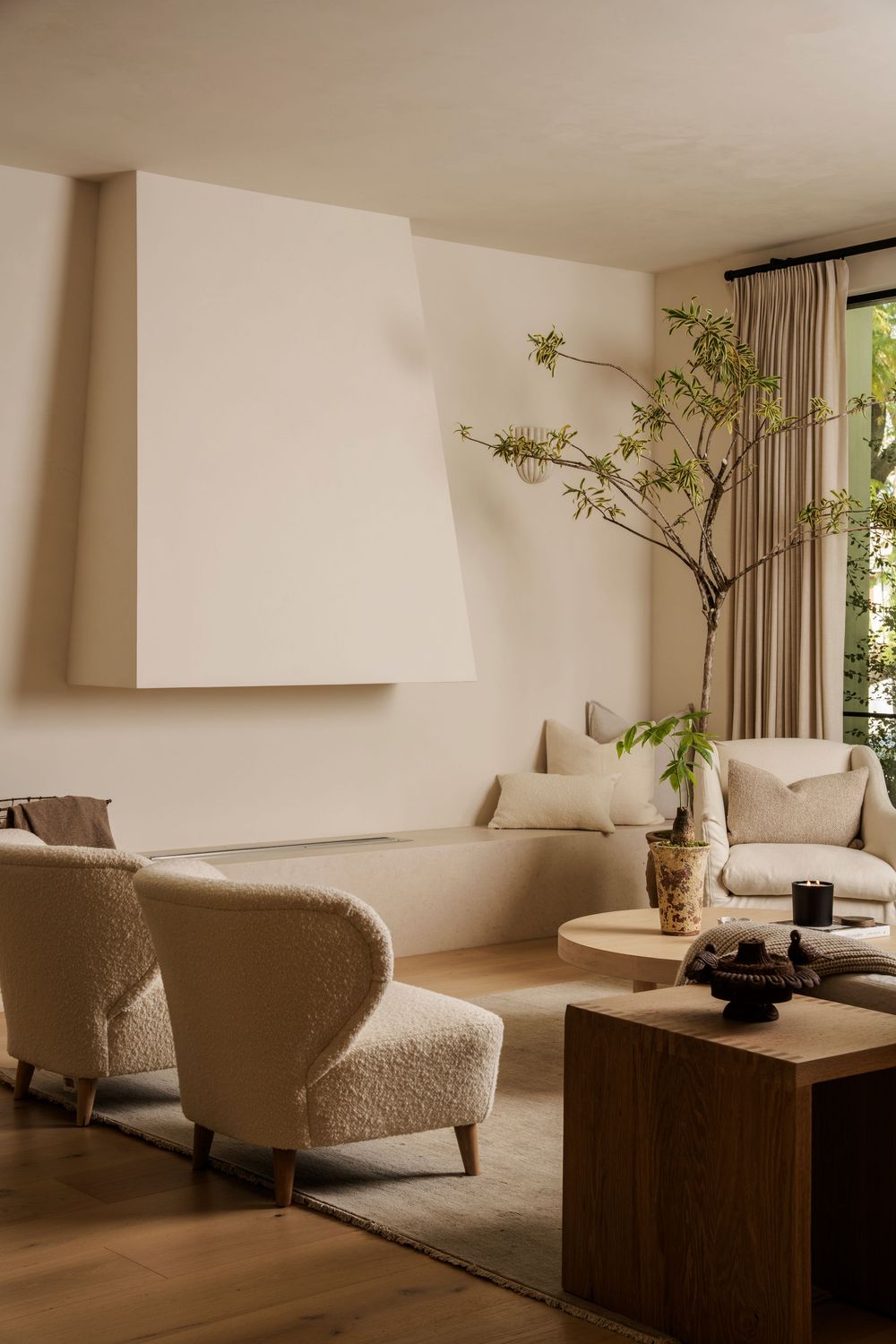 Minimalist living room with beige bouclé armchair, wooden coffee table, neutral sofa, and tall branch in vase by window.