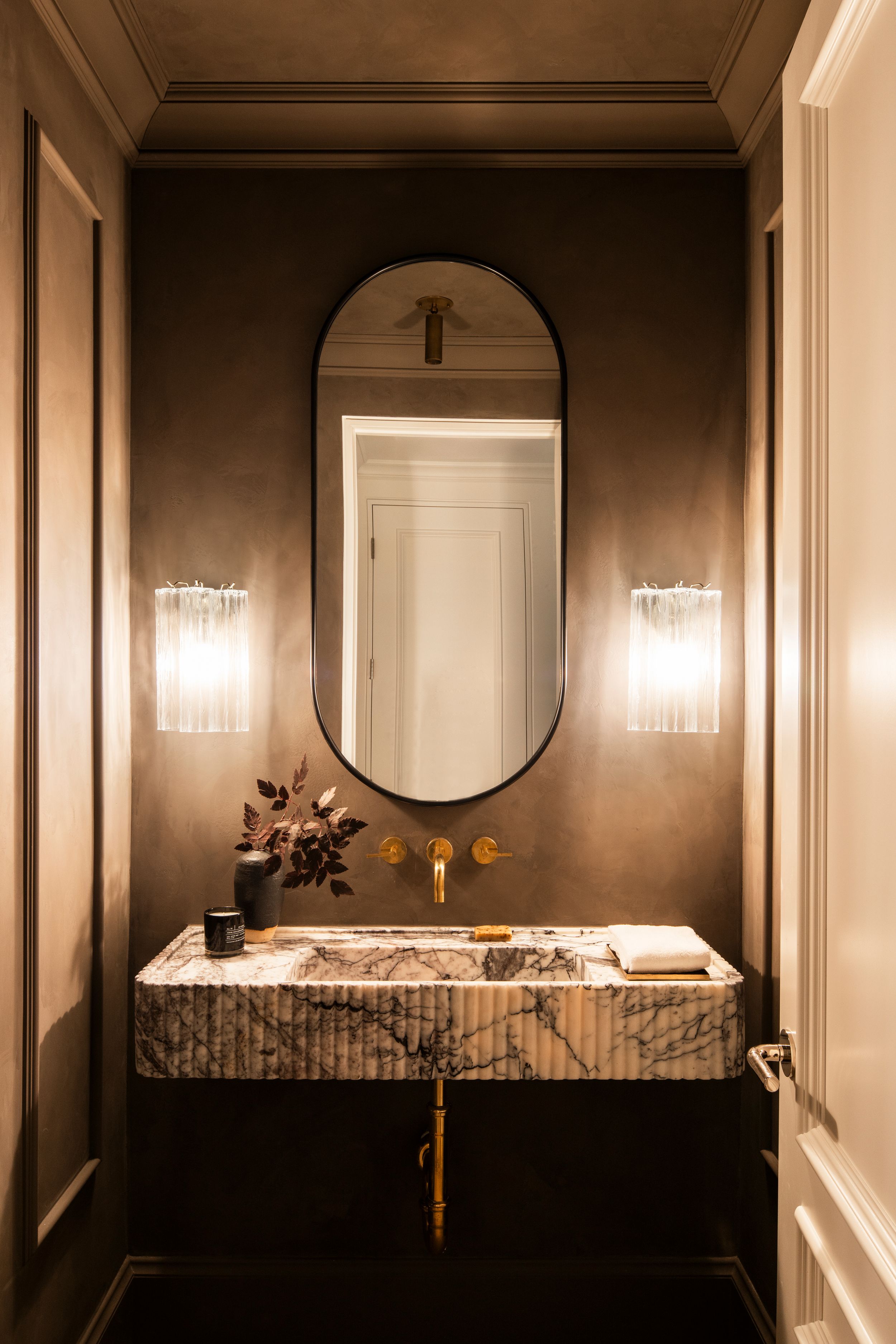 Modern bathroom interior with dark walls, round mirror, crystal sconces and white marble countertop.