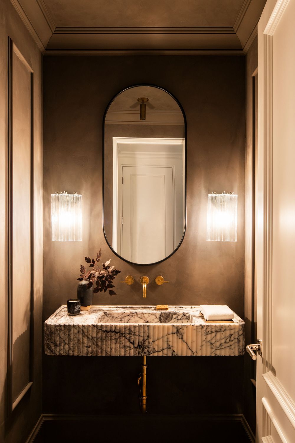 Modern bathroom interior with dark walls, round mirror, crystal sconces and white marble countertop.