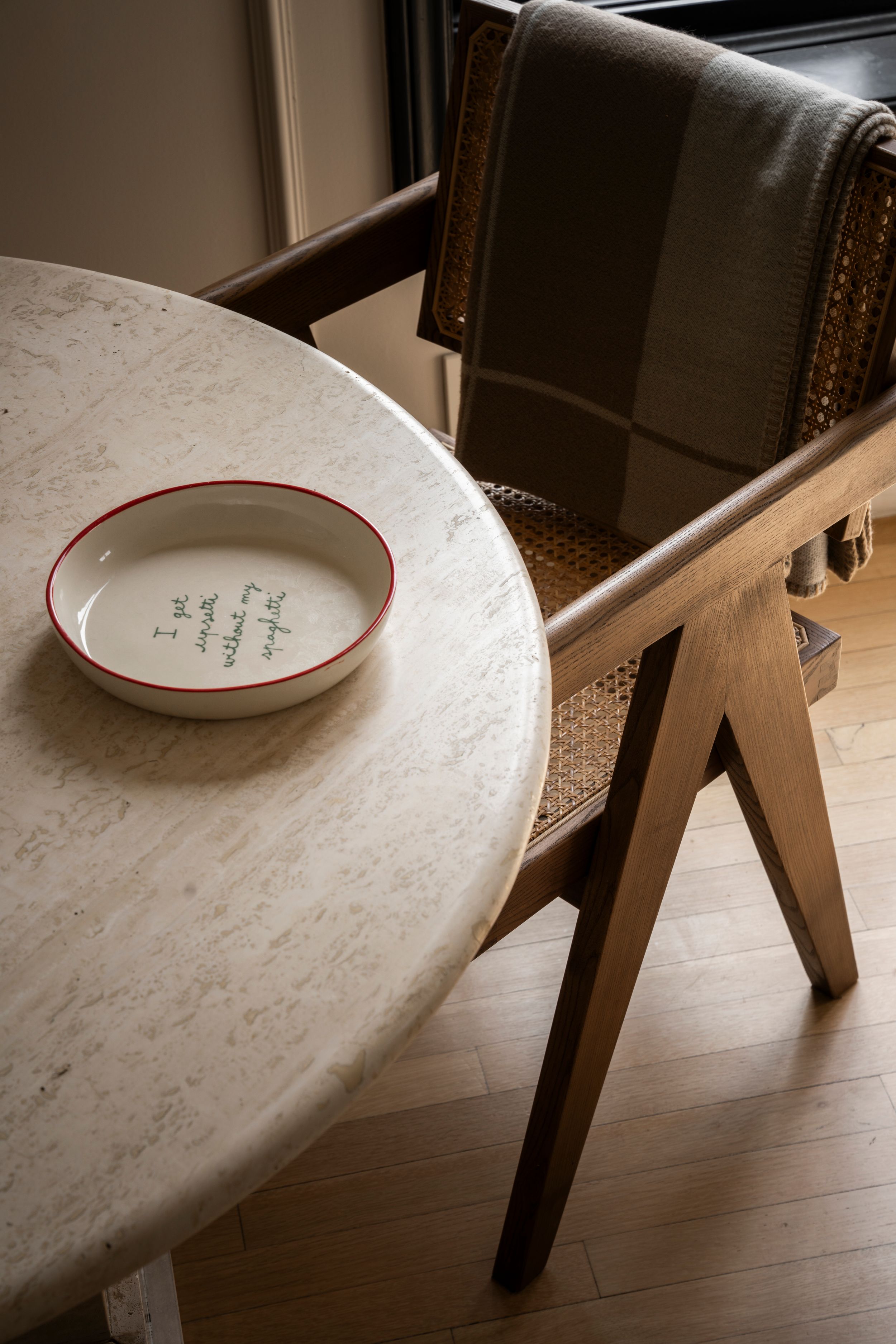 A white ceramic bowl with red rim and handwritten text sits on a round marble table beside a wooden rattan chair.