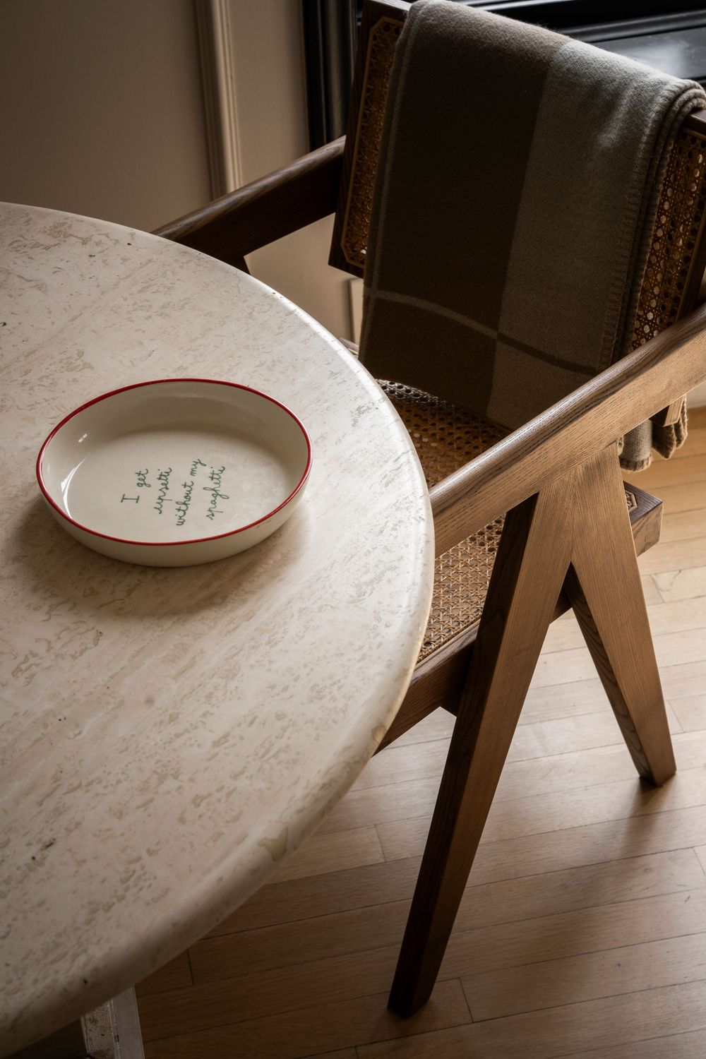 A white ceramic bowl with red rim and handwritten text sits on a round marble table beside a wooden rattan chair.