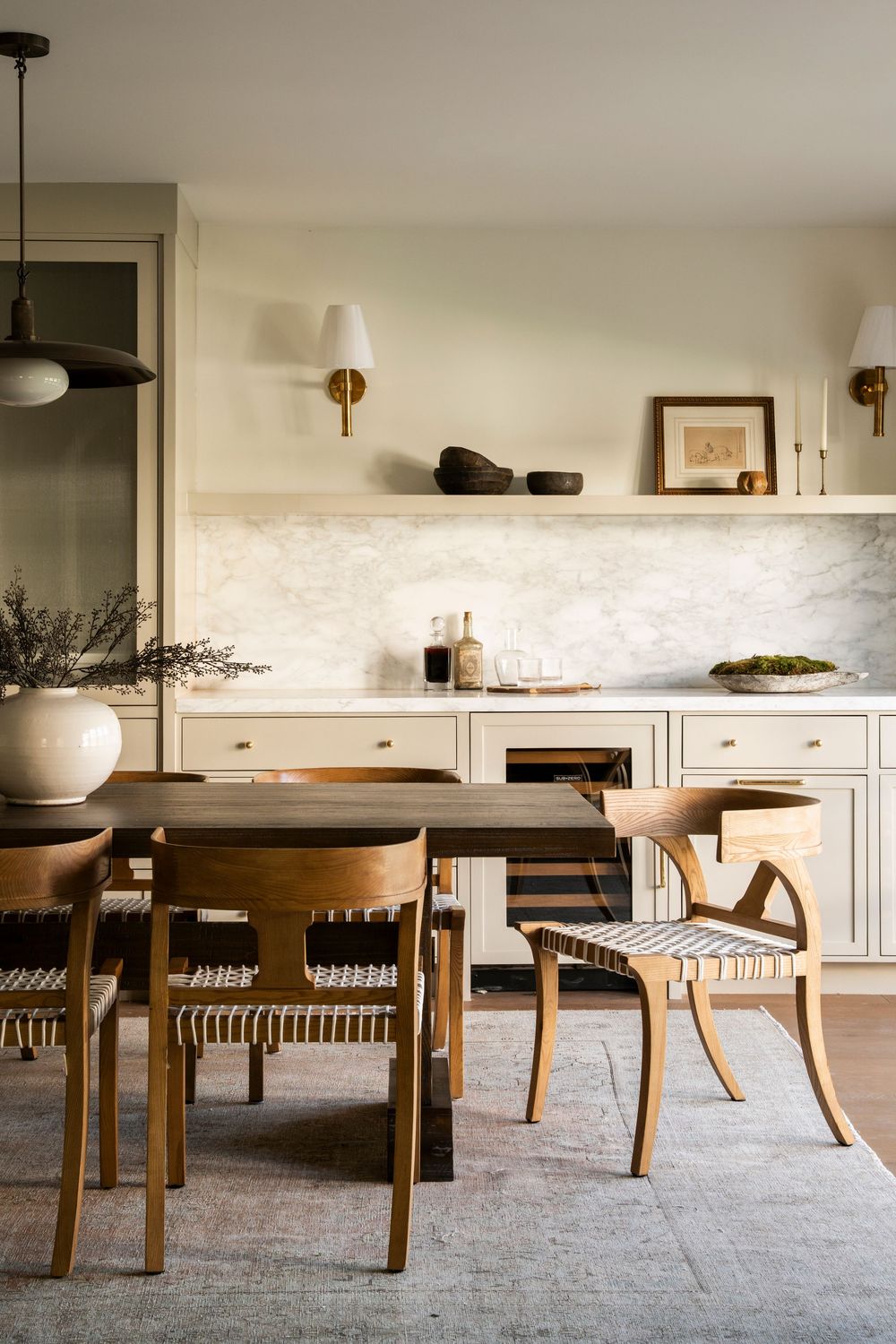 Warm dining room with wooden chairs, marble backsplash, cream cabinets, brass sconces, and a shelf with decorative objects.