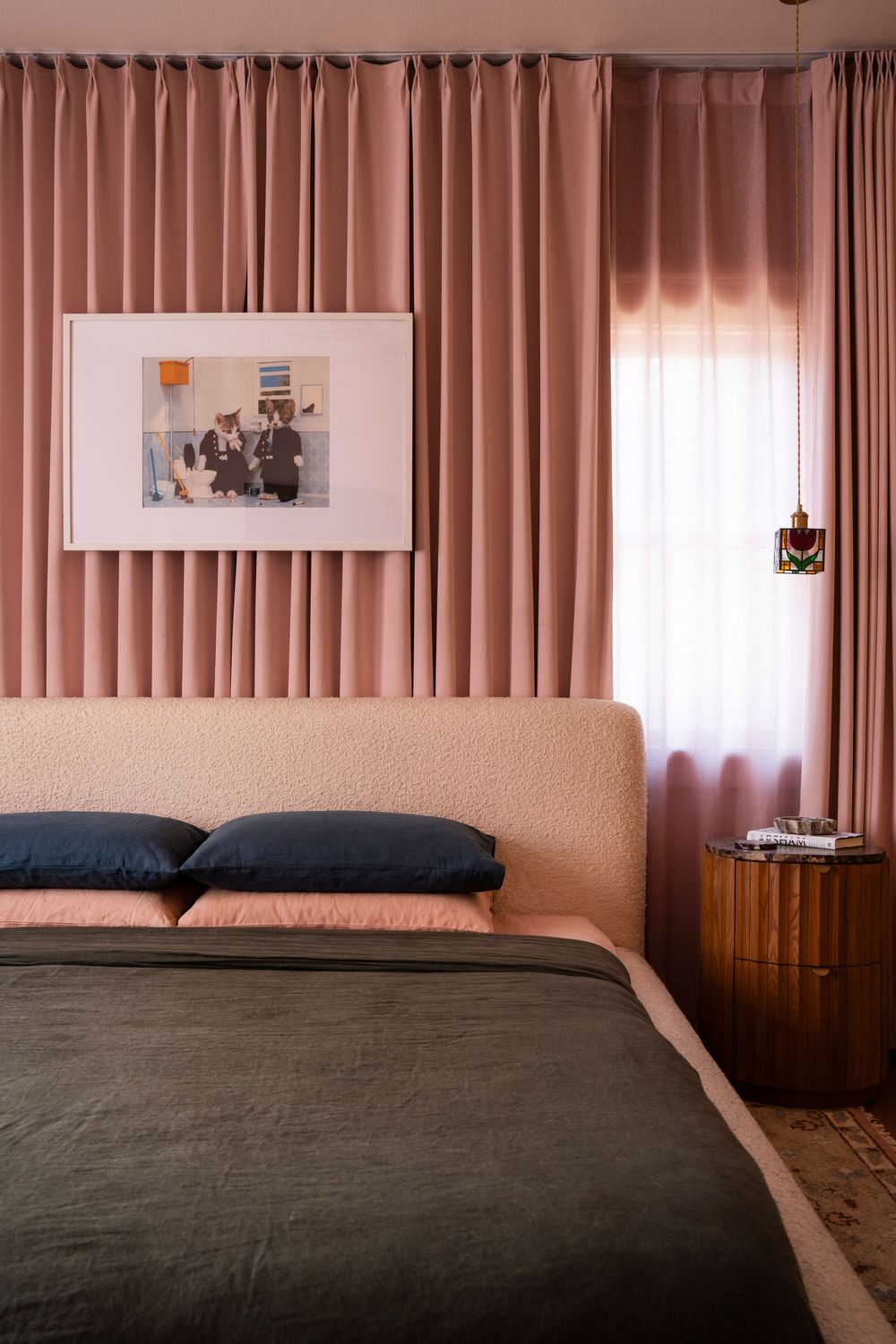 Pink draped bedroom with boucle headboard, dark bedding, walnut nightstand, framed artwork, and pendant light.