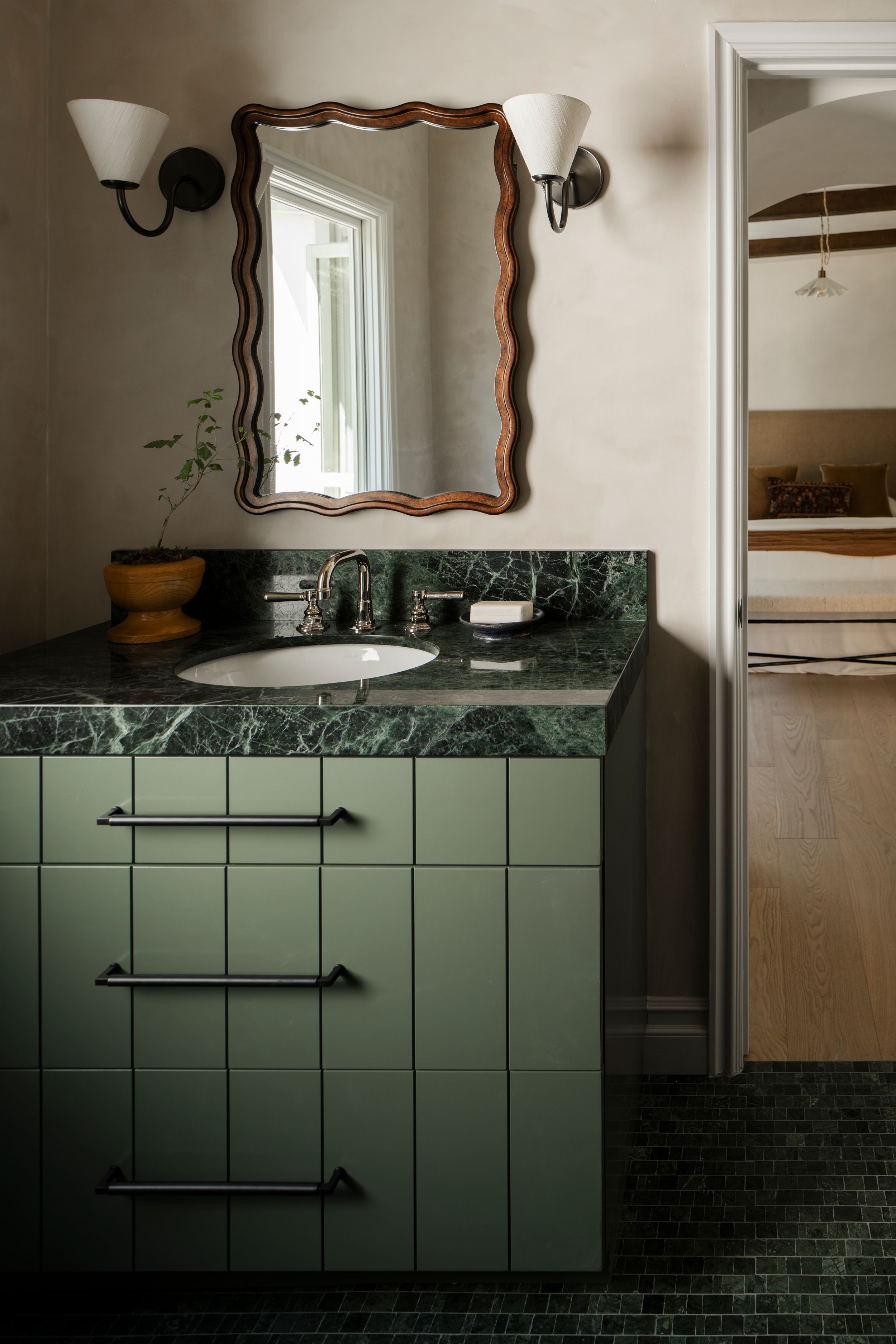 Green marble bathroom vanity with wavy mirror, sconce lighting, and dark green tile flooring in elegant bathroom.