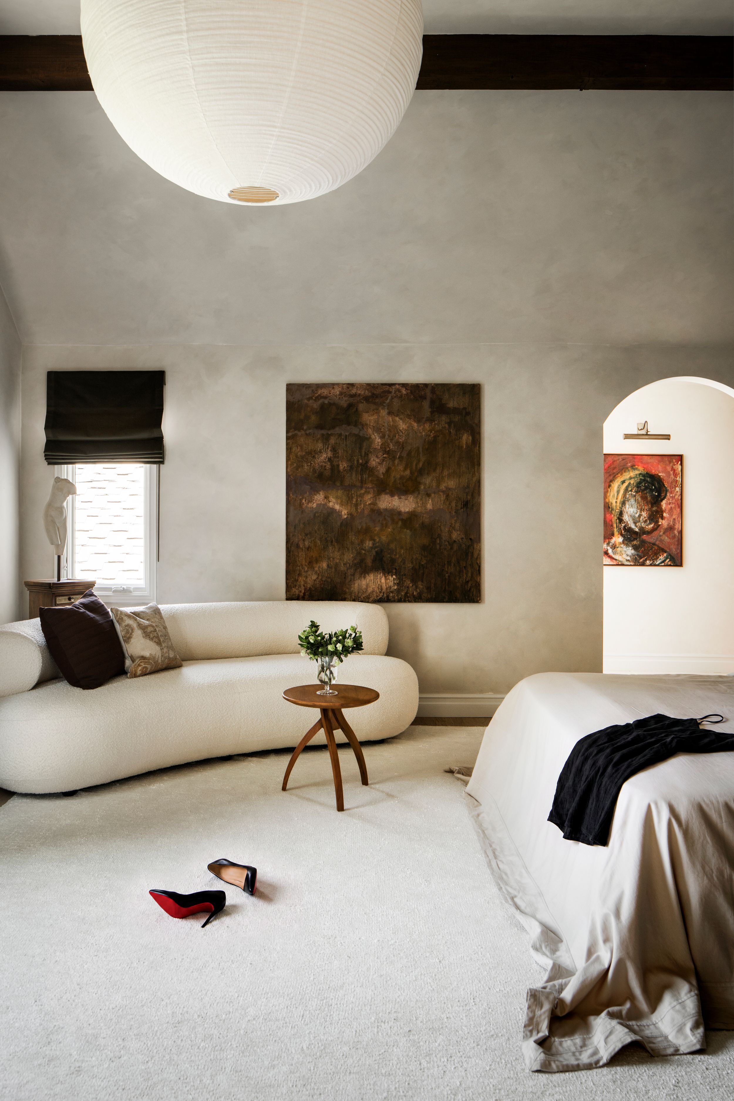 Minimalist bedroom with curved cream sofa, paper lantern pendant, abstract art, and red Louboutin heels on carpet.