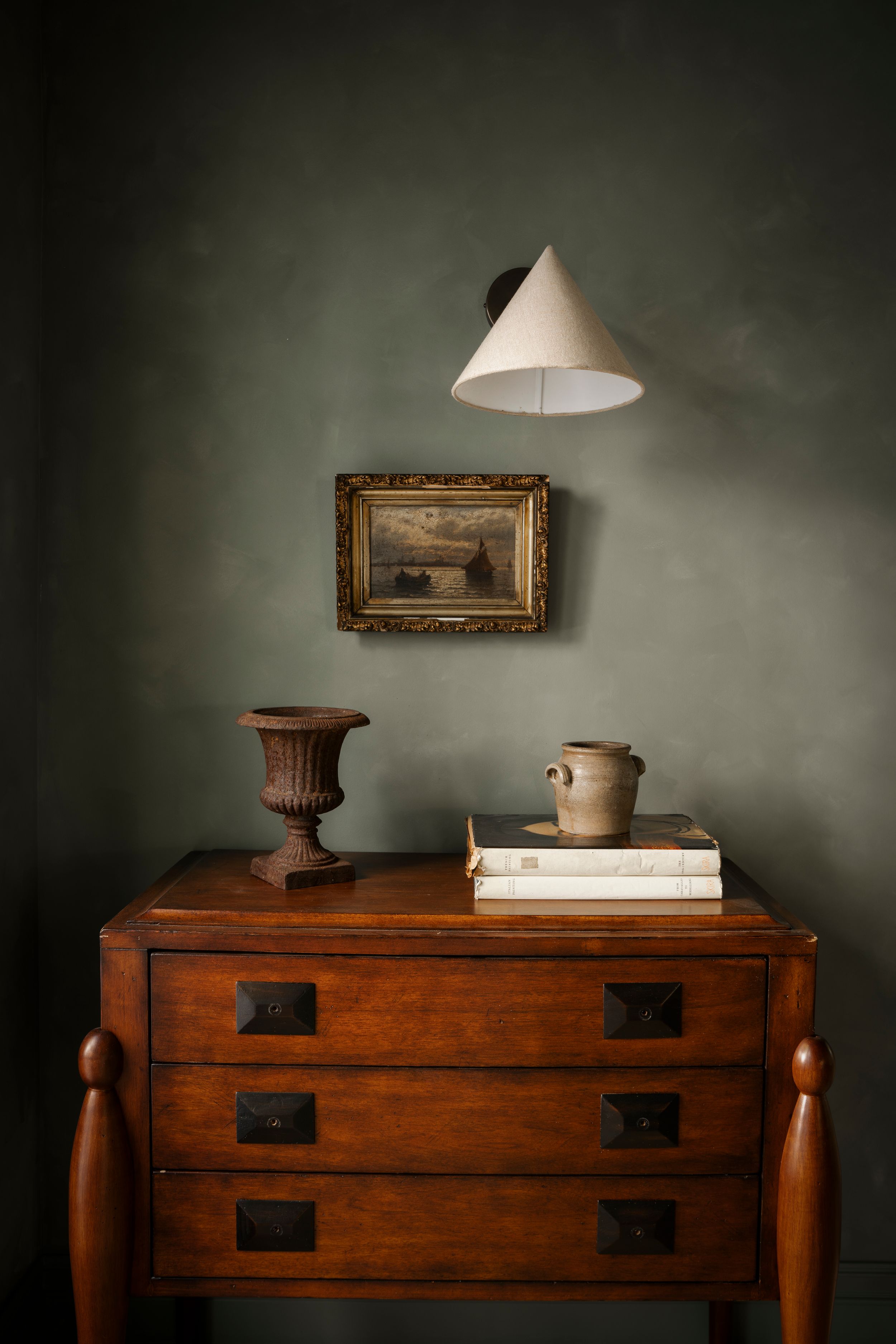 Vintage wooden dresser with books, ceramic mug, and urn vase beneath a wall lamp and gold-framed painting on sage green wall.
