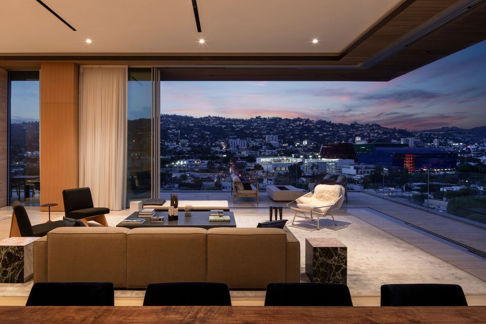 Luxury living room with panoramic city views at dusk, featuring modern furniture and an open terrace overlooking hillside homes.