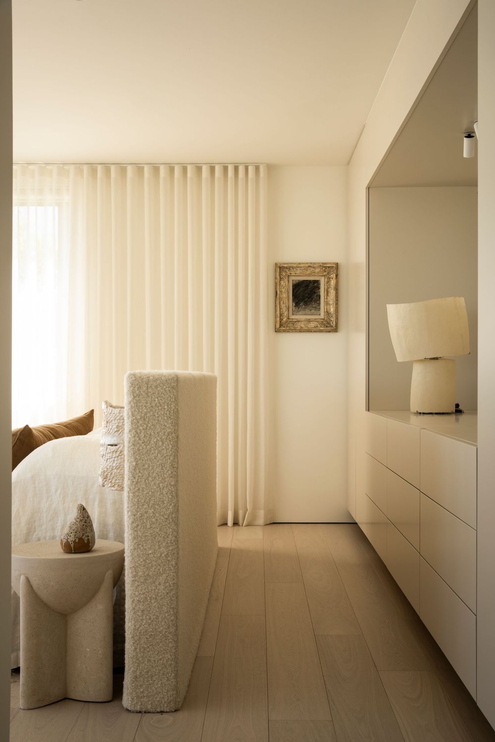 Minimalist bedroom with cream curtains, built-in white dresser, table lamp, and textured sculptural room divider on light wood floors.