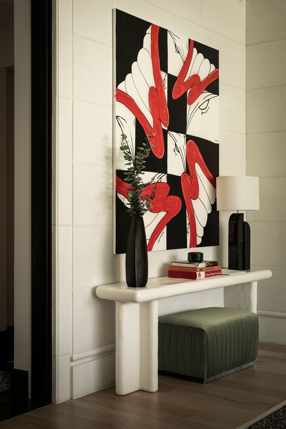 White console table with black vases, green ottoman, and bold red and black abstract artwork in an elegant entryway.