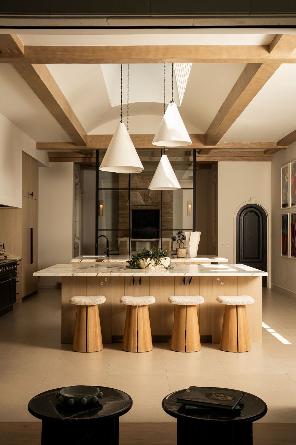 Modern kitchen with wooden beam ceiling, white pendant lights, marble island, and natural wood stools in warm neutral tones.