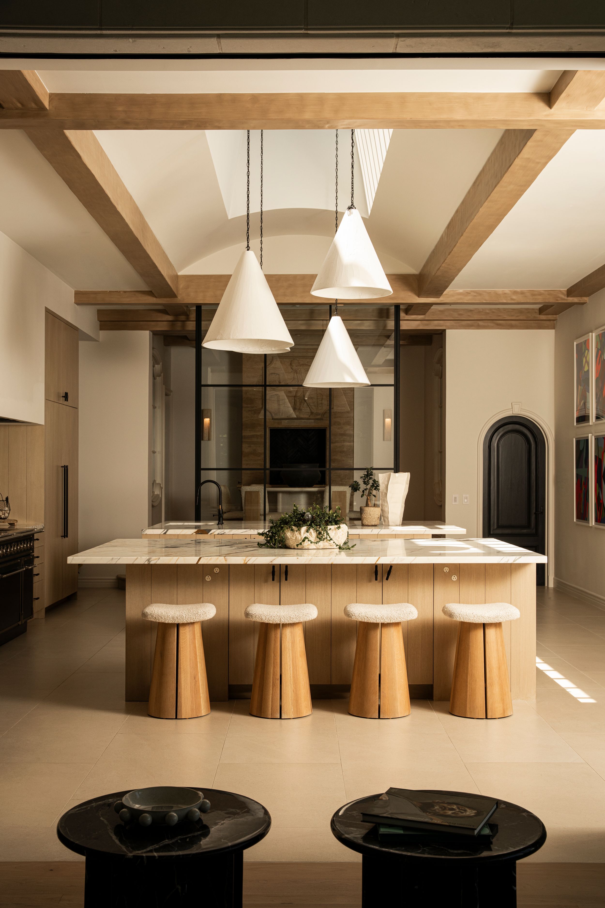 Modern kitchen with wooden beam ceiling, white pendant lights, marble island, and natural wood stools in warm neutral tones.