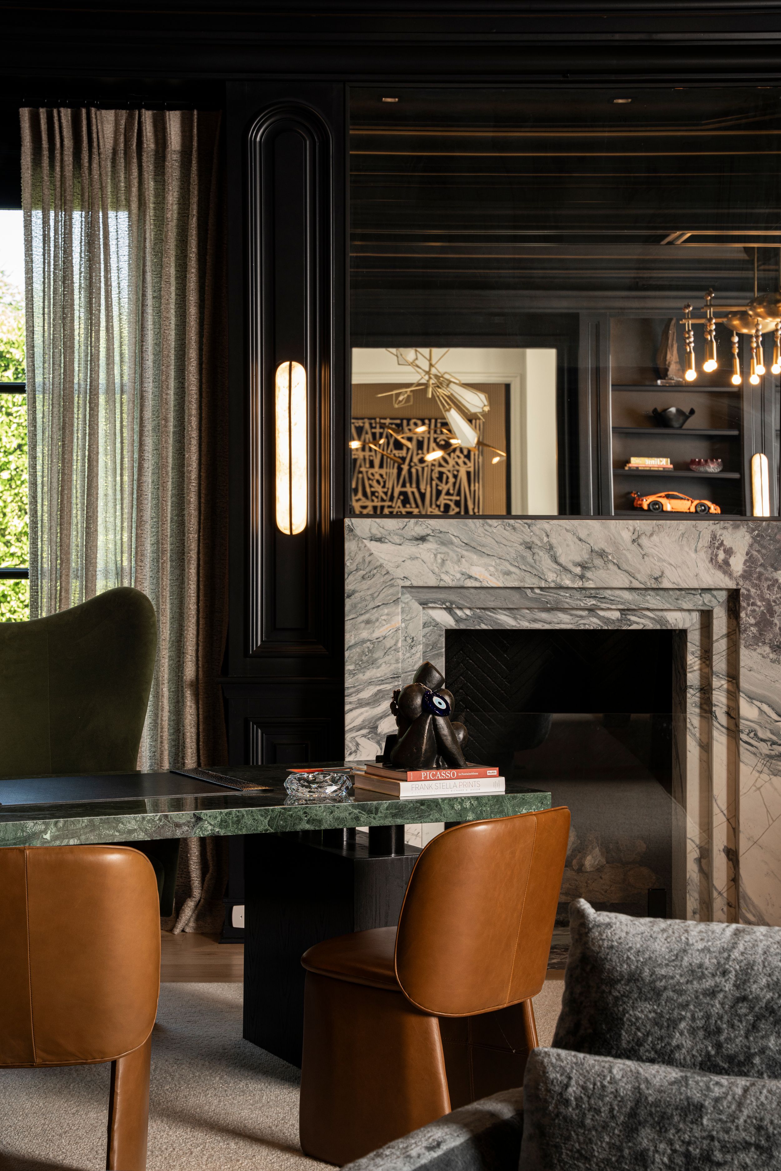 Elegant dark interior with marble fireplace, green dining table, tan leather chairs, and gold accents in a sophisticated living space.