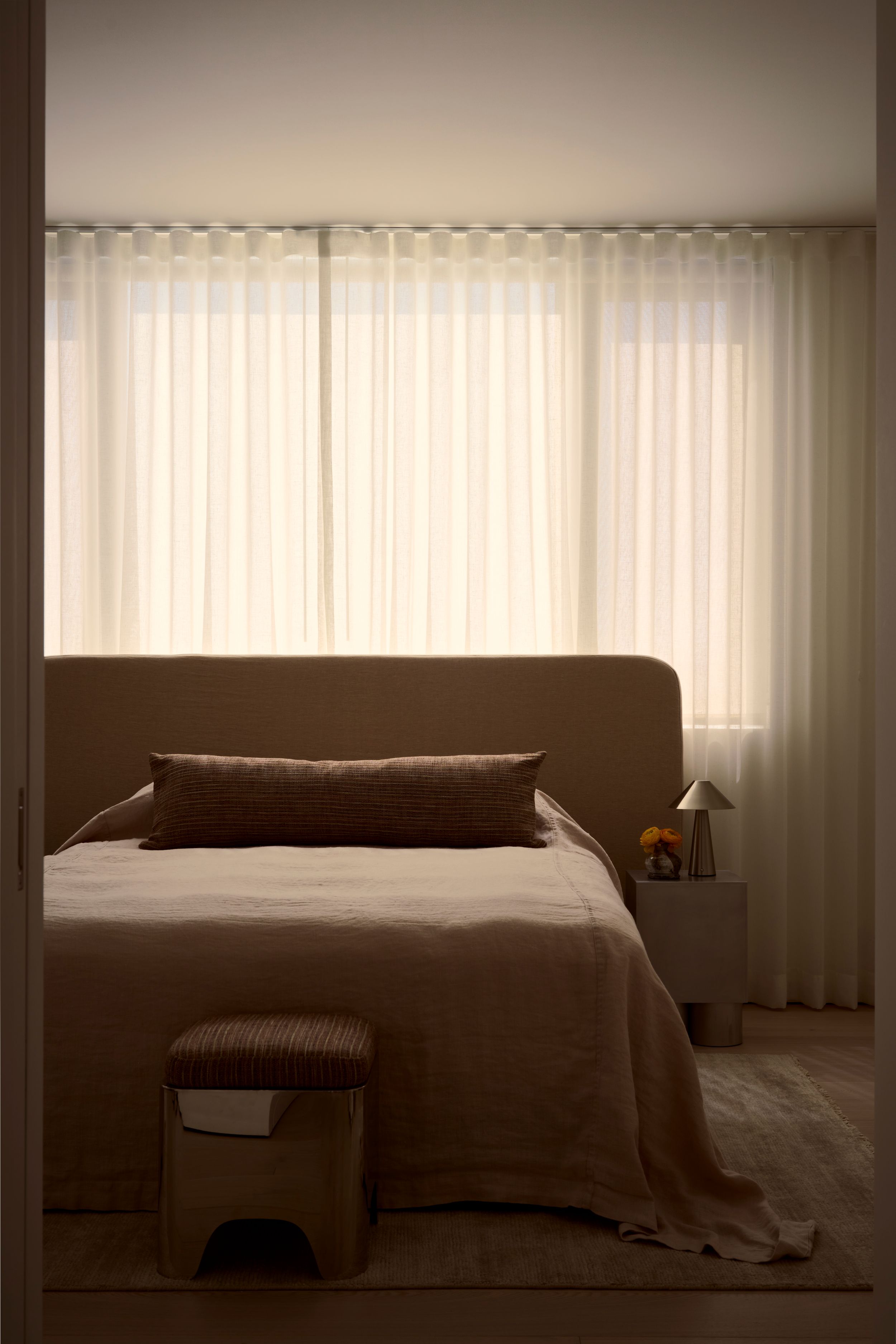 Minimalist bedroom with taupe upholstered bed, linen bedding, sheer curtains, and a small corduroy stool in warm neutral tones.