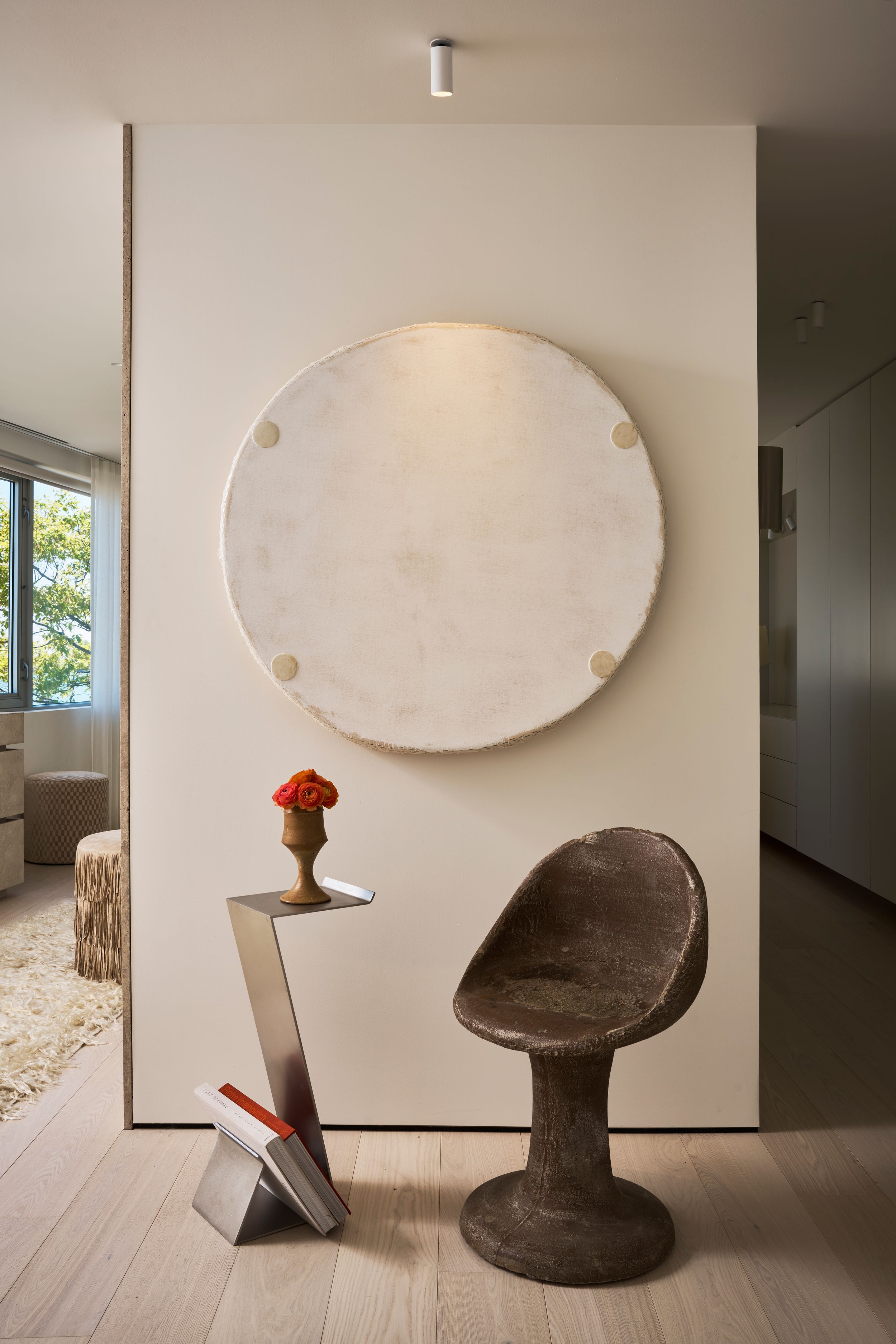 Modern interior with circular wall art, sculptural side table with red flower, and a textured brown chair on light wood floor.