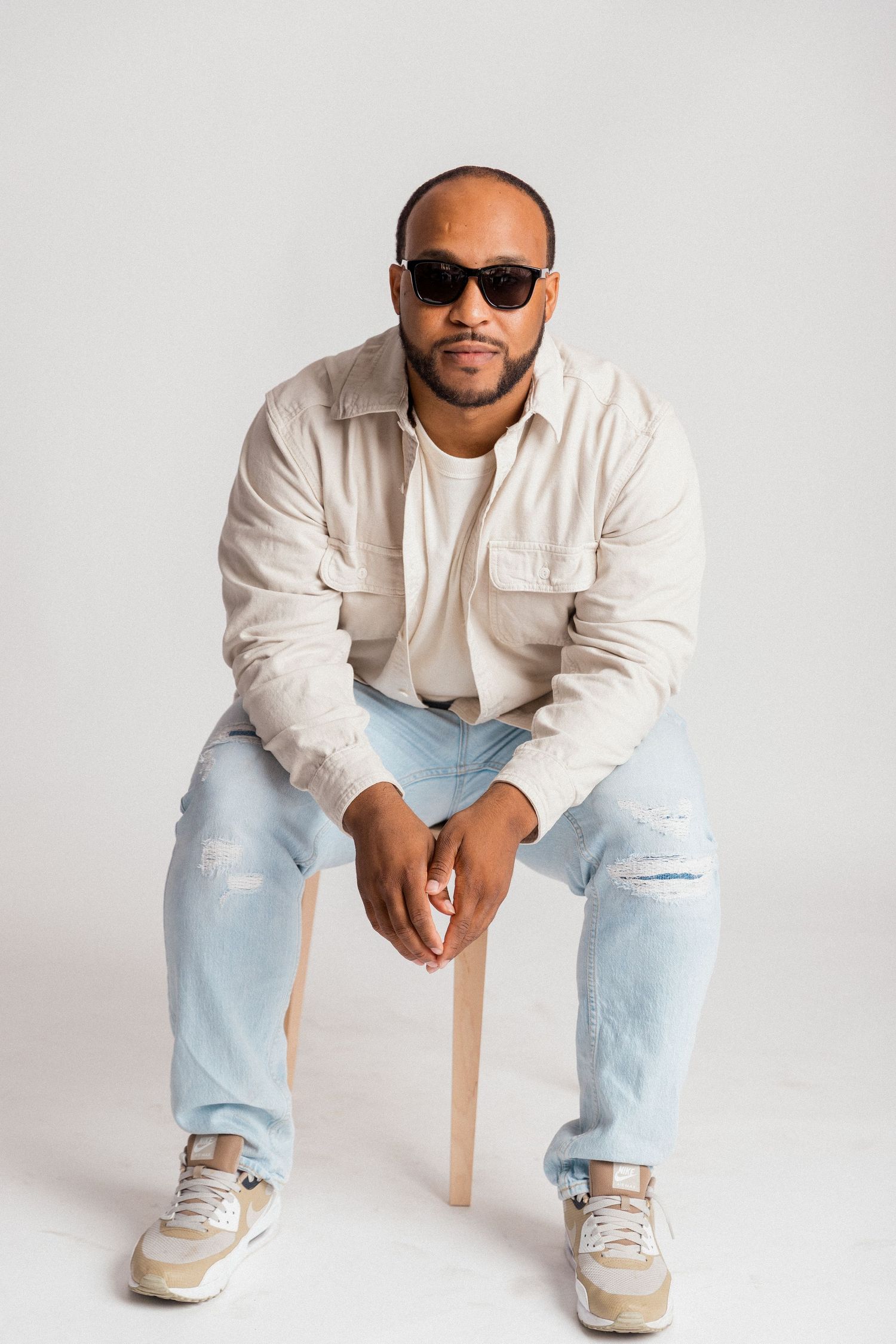 Stylish photographer in white shirt and jeans holding camera against white backdrop.