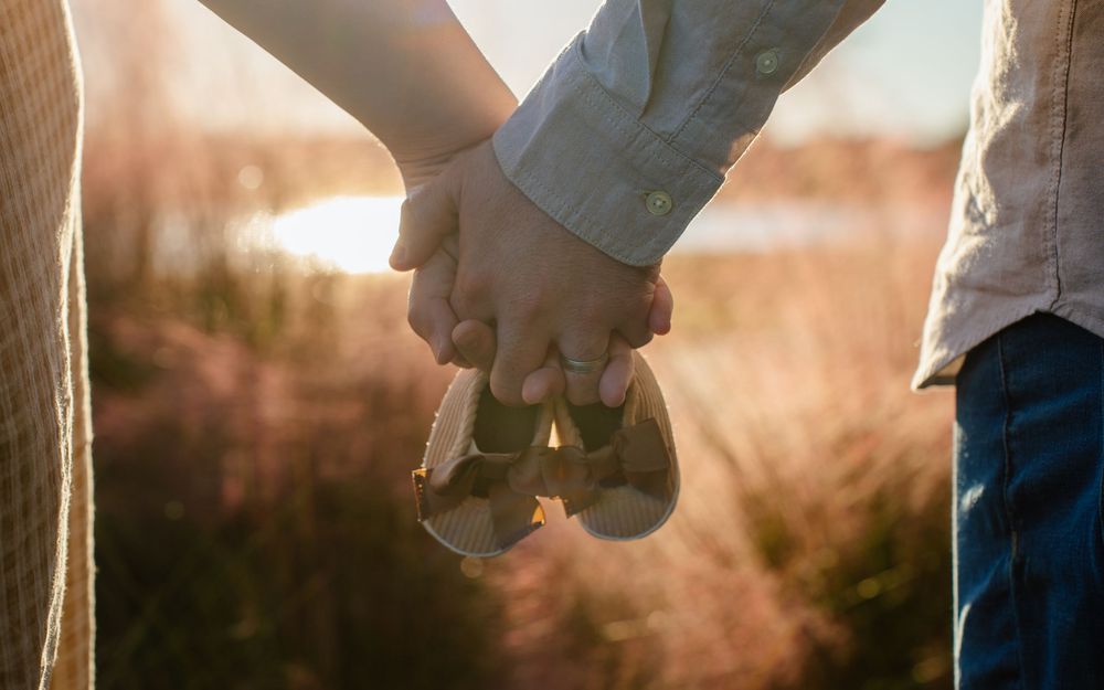Couple holding hands and baby shoes in a golden field, maternity announcement photo.