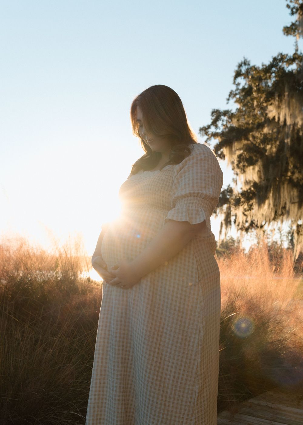 Pregnant woman in gingham dress holding baby bump at golden hour sunset outdoors.