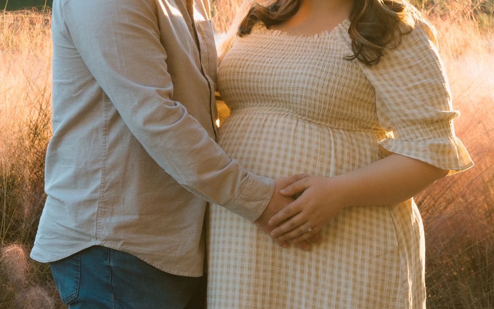 Pregnant woman in a gingham dress with partner outdoors during golden hour maternity photoshoot.