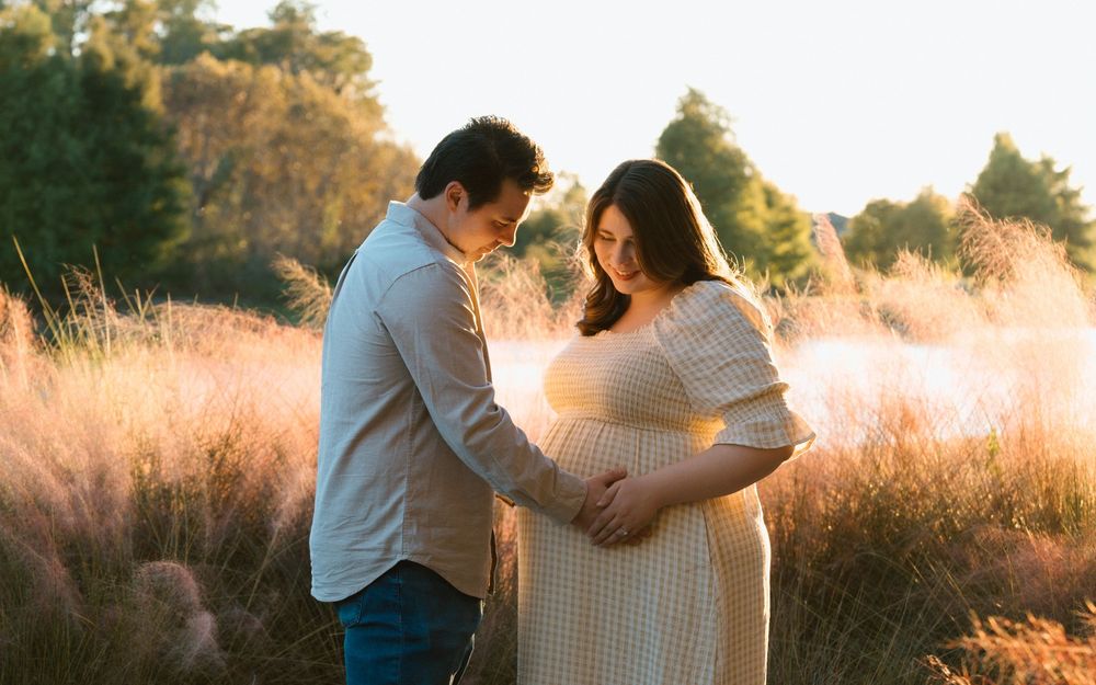 Couple tenderly cradling pregnant belly in golden sunset field with tall grasses and warm bokeh background.
