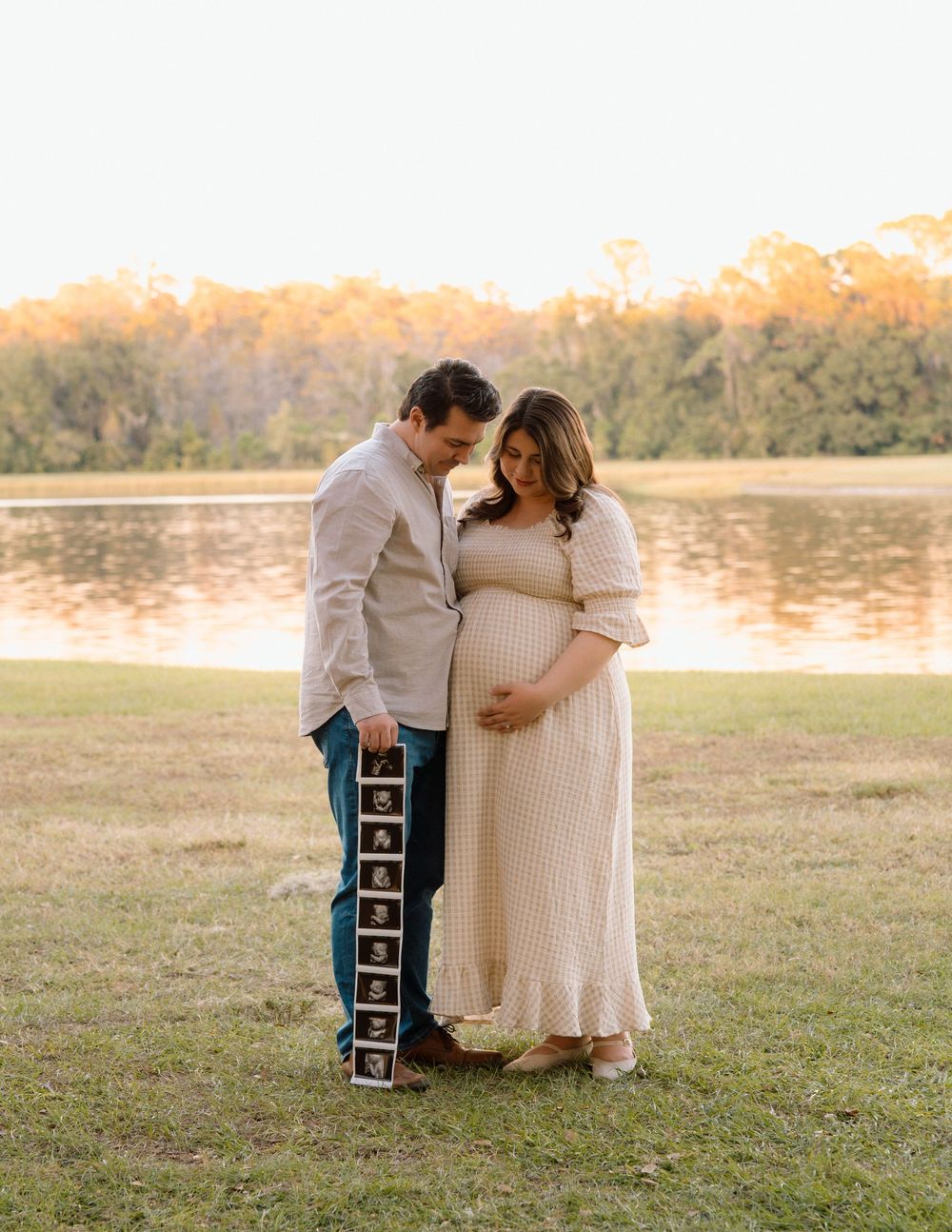 Couple posing for maternity photo by a lake at golden hour, woman in cream dress cradling baby bump.