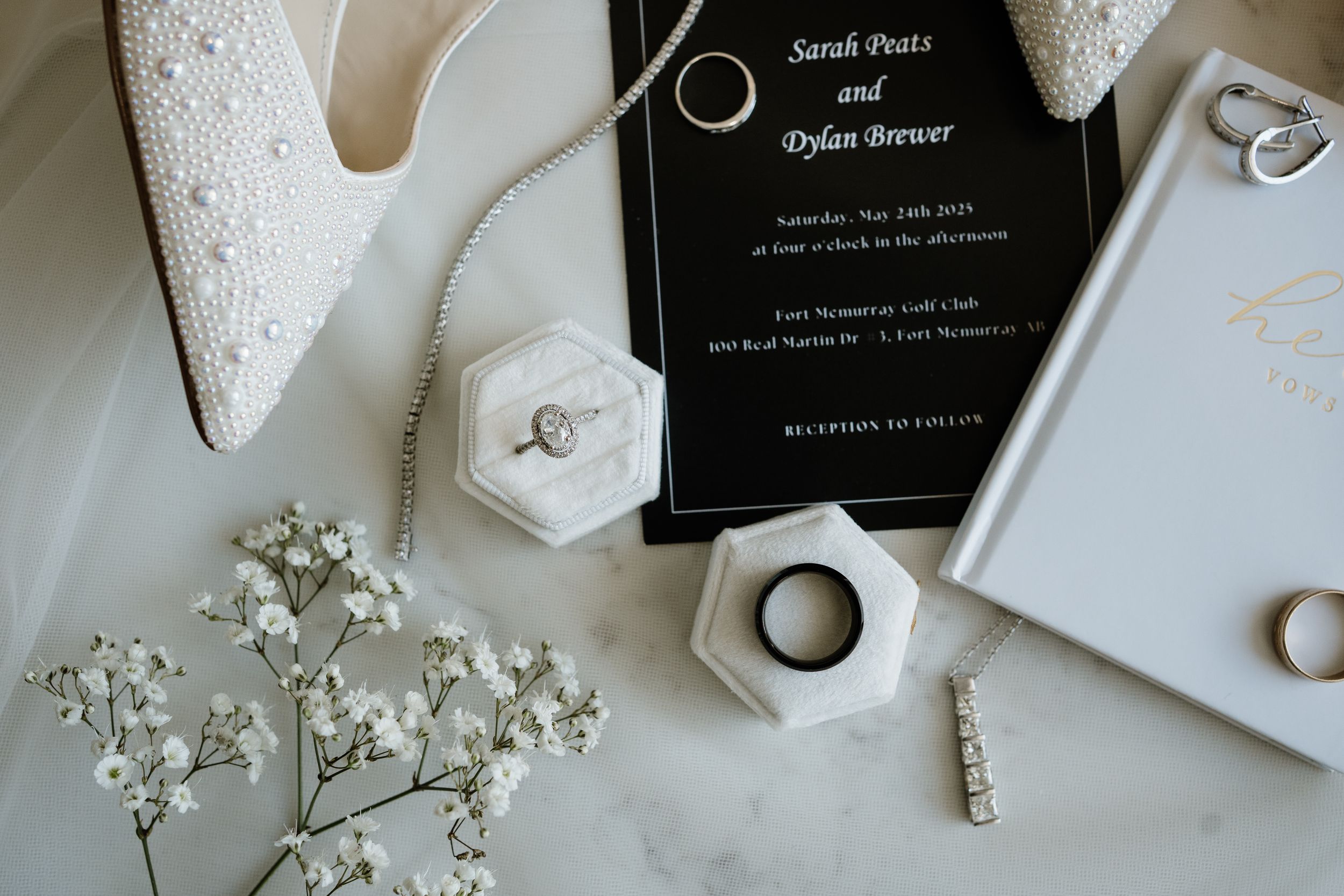 Wedding details flatlay with white ring boxes, baby's breath flowers, invitation card, and bridal shoes on marble surface.