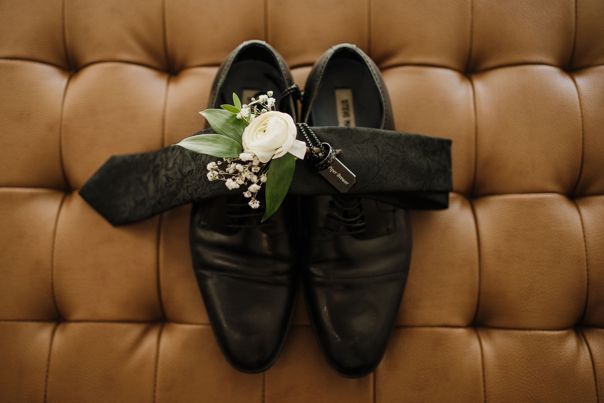 Black leather dress shoes with white rose and baby's breath boutonniere on brown tufted leather background.