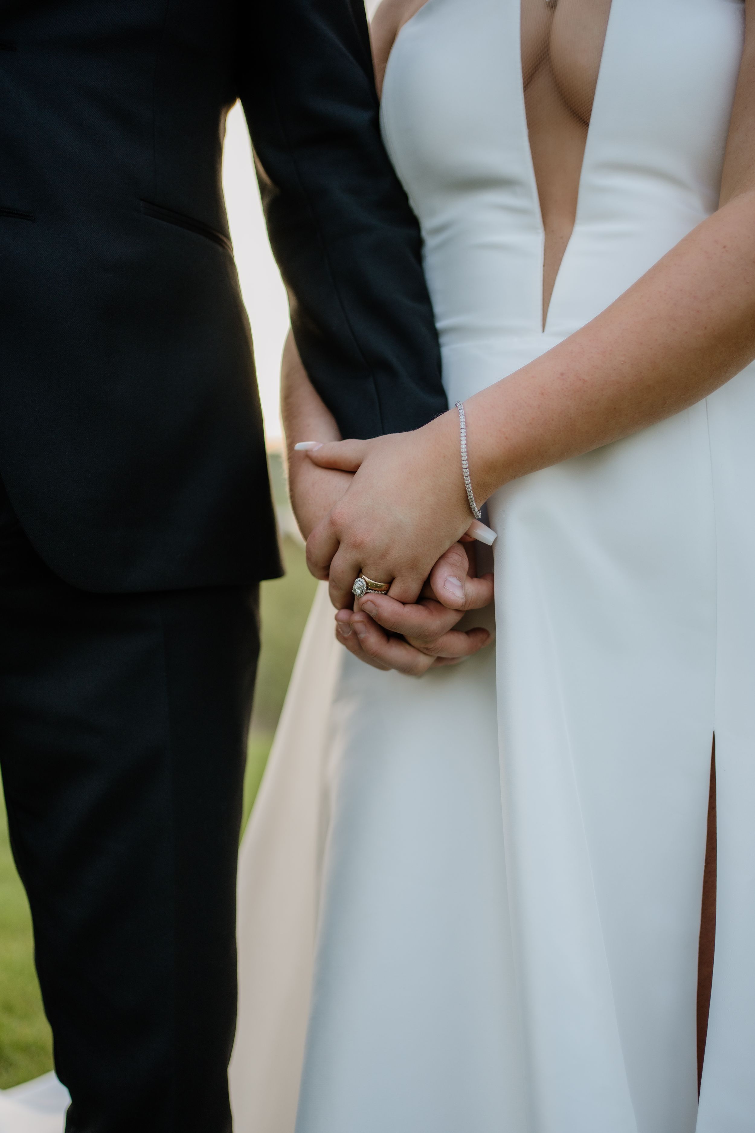 Close-up of couple holding hands at wedding, showing elegant white bridal gown and black suit sleeve details.
