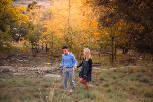 A couple walks together through golden autumn trees on a grassy hillside during sunset.