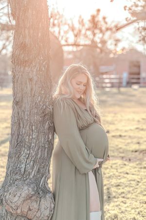 Silhouetted against golden sunlight, an expectant mother in a flowing dress cradles her baby bump beside a tree at sunset.