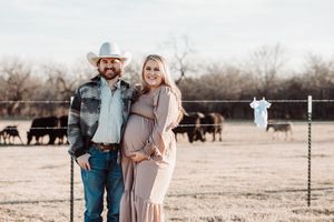 A couple poses together on a ranch with cattle grazing in the background, expecting a new addition to their family.