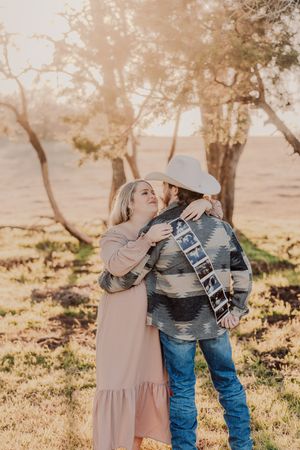 Couple embraces in a romantic sunset moment under trees wearing western attire and cowboy hat.
