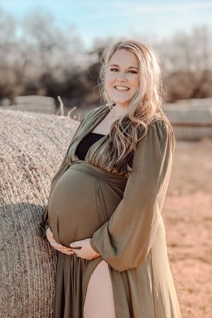 Expecting mother in olive green dress poses against hay bale outdoors in warm sunlit setting.