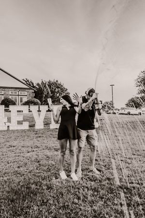 Two people in casual attire stand together in a grassy field on a sunny day, raising their arms in joyful celebration.