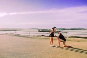 A romantic proposal takes place on a scenic hilltop during sunset with a beautiful ocean view in the background.