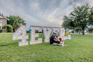 Decorative white letters spelling 'TEXAS' sit on a green lawn with someone sitting beside them on a sunny day.