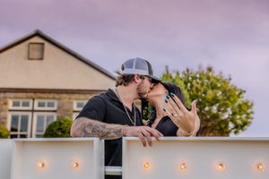 Two people share a romantic kiss at sunset while leaning against a white wall decorated with glowing string lights.
