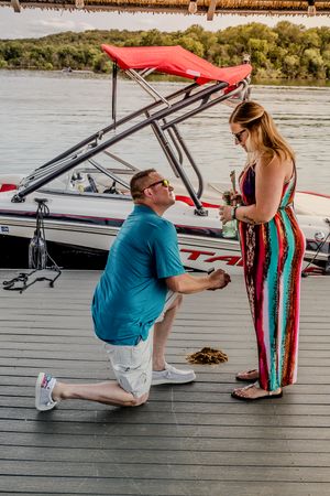 A romantic proposal taking place on a boat dock with a red wakeboarding boat visible in the background.