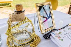 Decorative gold beads and a small bottle alongside a framed photo and event invitation card on a white tablecloth.