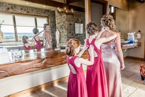 Wedding party members wearing pink and rose-colored dresses prepare in a rustic room with stone walls and large windows.